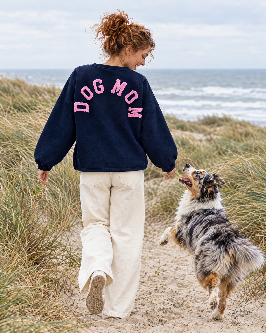 Frau im Dog Mom Sweatshirt in Navy mit rosa Schrift und Australian Shepherd in den Dünen (KI-Bild)