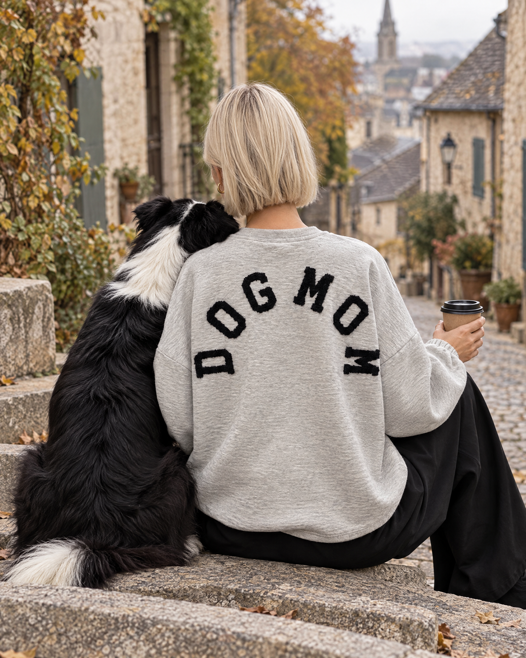 Frau im Dog Mom Sweatshirt in Hellgrau mit schwarzer Schrift und Border Collie in der Altstadt (KI-Bild)